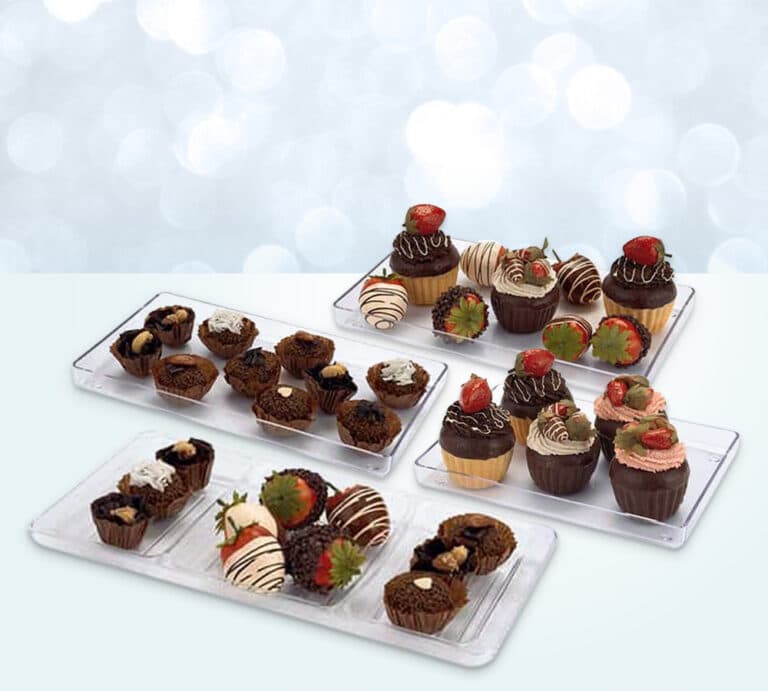 A selection of assorted desserts including chocolate-covered strawberries, cupcakes, and small pastries arranged on clear candy trays. The trays are displayed on a light blue surface with a blurred bokeh background, creating an inviting and festive atmosphere. These candy trays are made by Pioneer